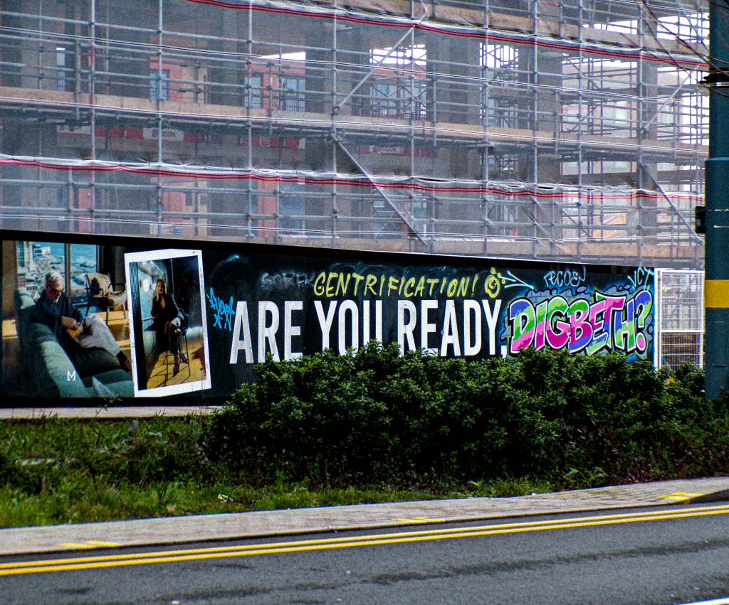 a sign in front of a high rise development site saying 'are you ready digbeth?' with a grafitti sign saying 'gentrification'